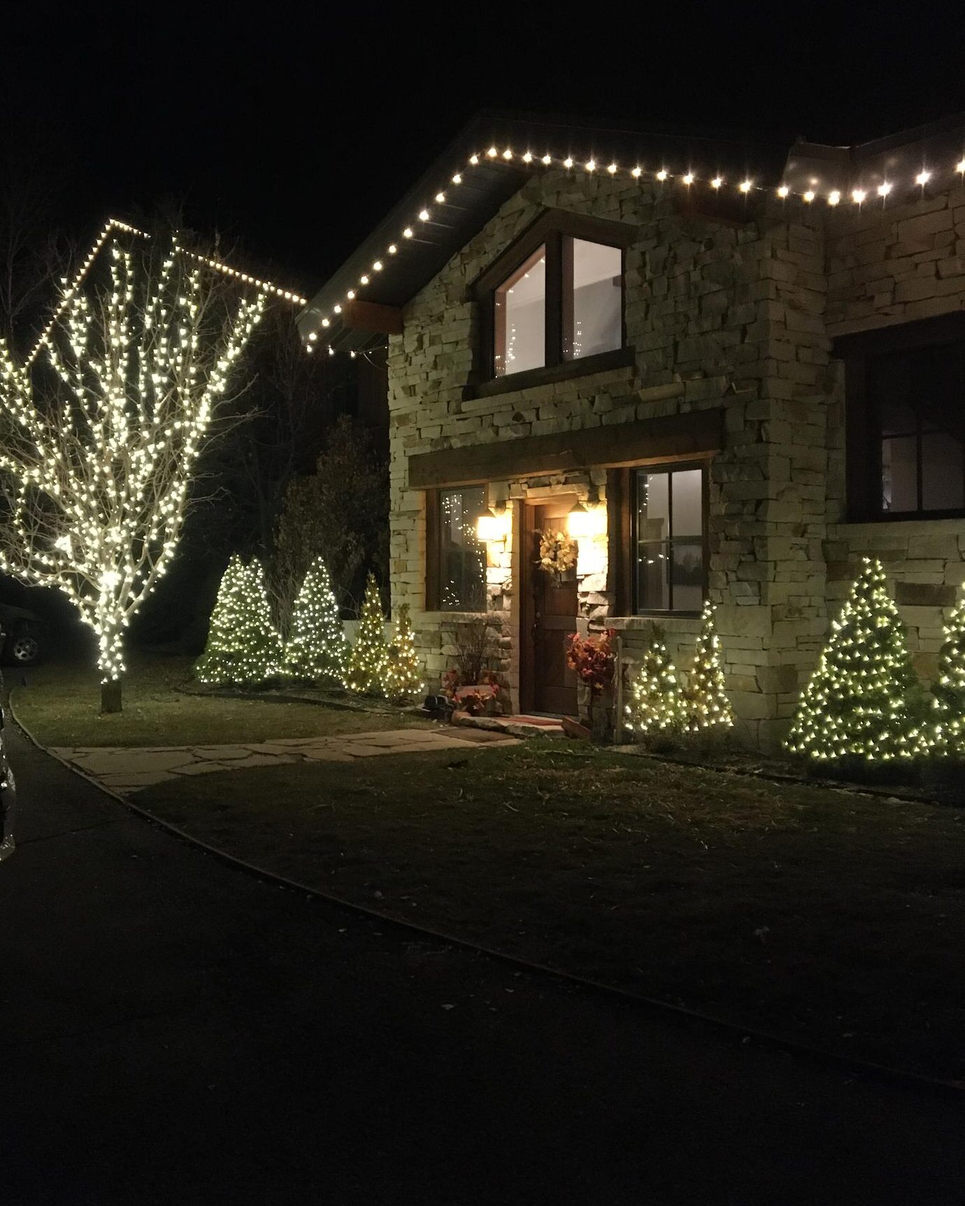 Stone house with roofline lights and a wrapped tree at night